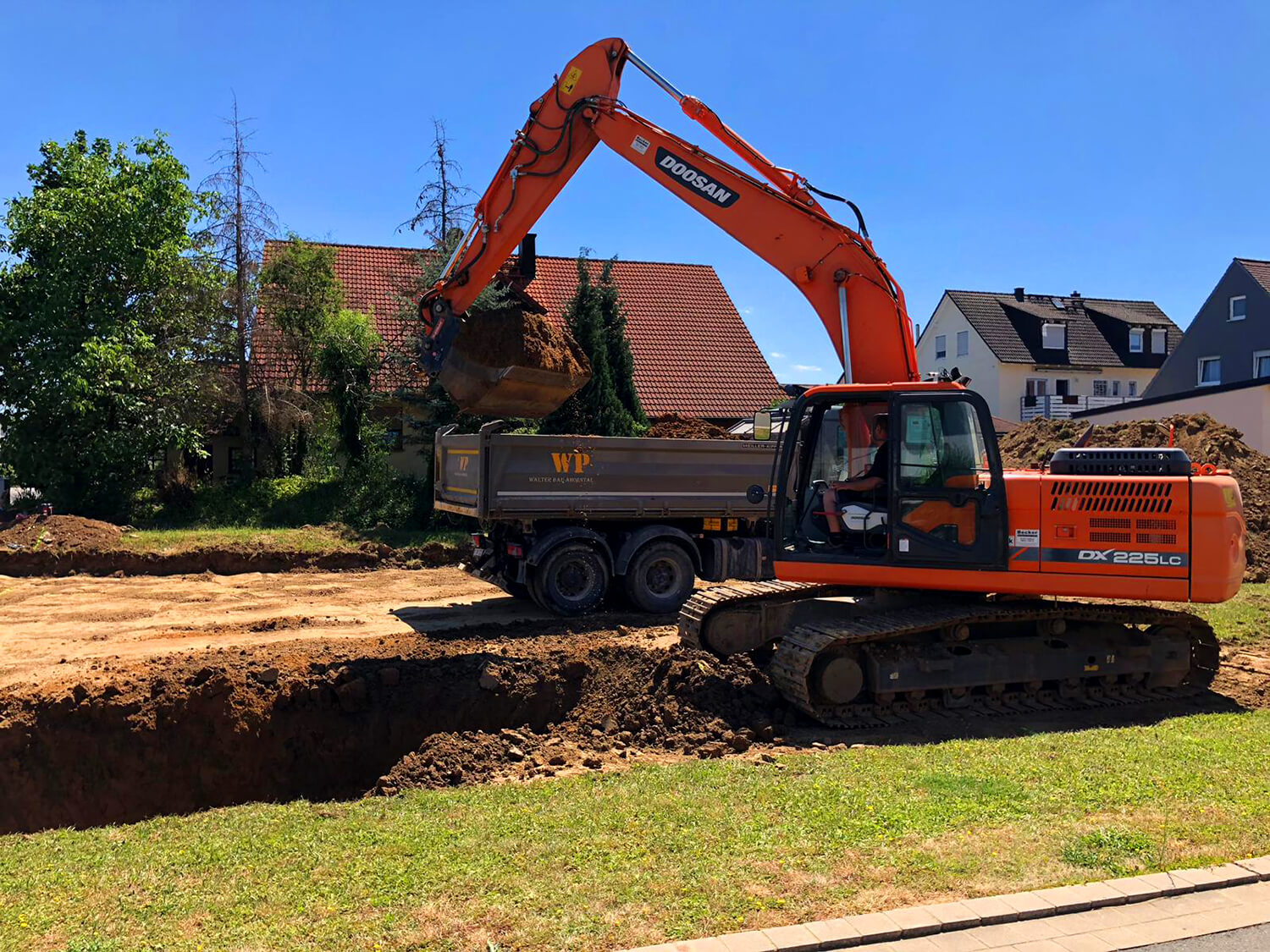 Kettenbagger baggert Kanal für Anschlüsse, LKW transportiert Erdaushub ab